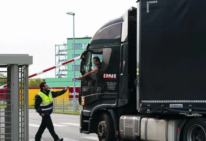 ADU Sicherheitsdienst Mitarbeiter kontrolliert LKW Fahrer, der vor einen Hofeinfahrt wartet
